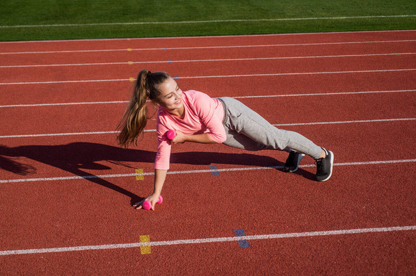 Power and balance. healthy childhood. workout on fresh air outdoor. confident athlete with barbell. teen girl warming up on stadium. kid in sportswear use dumbbell. child do exercise on racing track - Photo, image