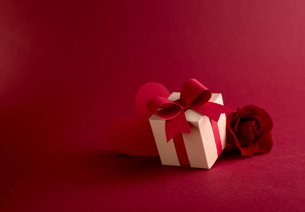 Happy St. Valentines day concept with craft paper gift box, red paper hearts and fresh rose against dark red background with empty space for text. - Фото, изображение