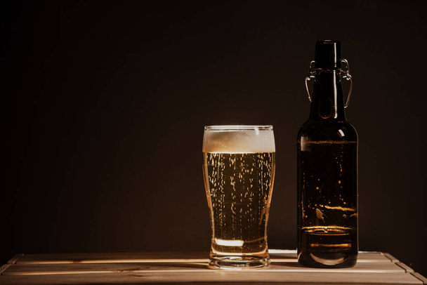 γεμάτο μπουκάλι μπύρας σε ξύλινο τραπέζι και ένα ποτήρι μπύρα σε μαύρο φόντο - Φωτογραφία, εικόνα