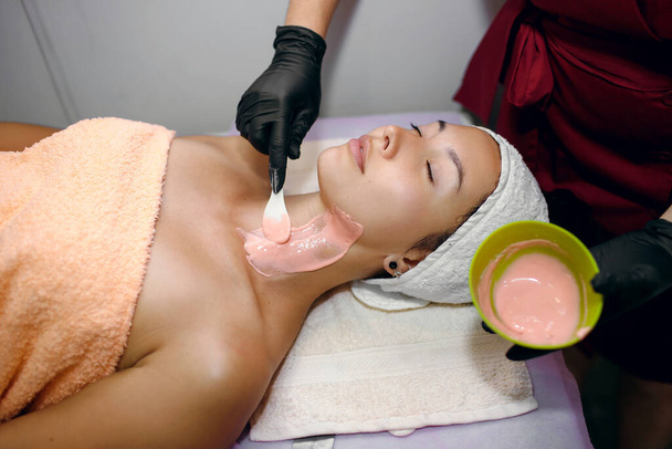 Uma menina bonita passa por procedimentos em um salão de spa, um esteticista mestre em luvas pretas aplica uma máscara rosa com uma camada espessa, limpando a pele da sujeira, mantendo um olho na pureza de seu rosto - Foto, Imagem