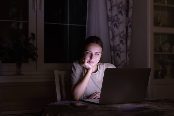 Γυναίκα κάθεται στο τραπέζι, βλέποντας μια ταινία στο φορητό υπολογιστή αργά το βράδυ στο σπίτι, μοιράζονται τα μέσα κοινωνικής δικτύωσης. - Φωτογραφία, εικόνα