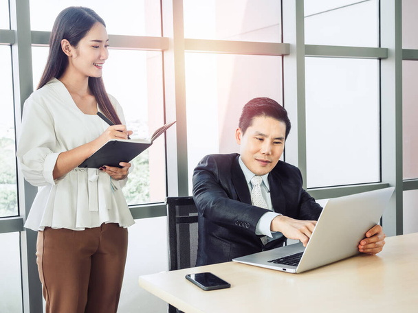 Ázsiai üzletember, öltönyös menedzser, aki a laptop képernyőjére mutat, és fiatal, mosolygó női titkárnő, akik együtt dolgoznak a sikerért. Munkatársak együttműködése a munkahelyen. - Fotó, kép