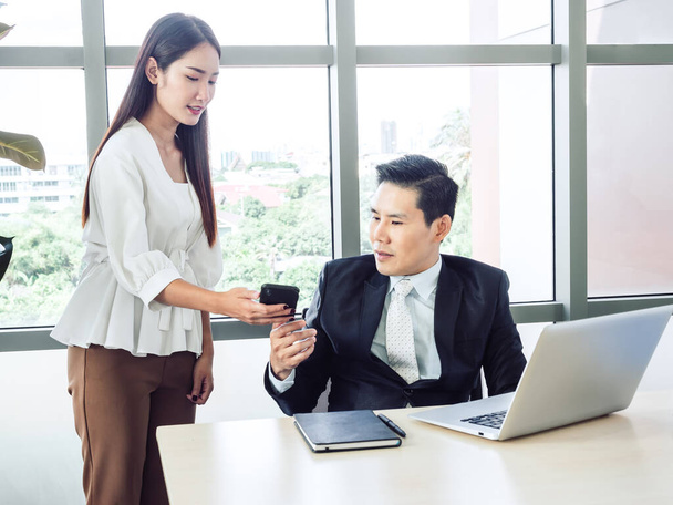 Azjatycki biznesmen, menedżer w garniturze pracujący z laptopem patrzący na smartfona od młodej sekretarki w biurze, pracujący razem dla sukcesu. Współpraca kolegów w miejscu pracy. - Zdjęcie, obraz
