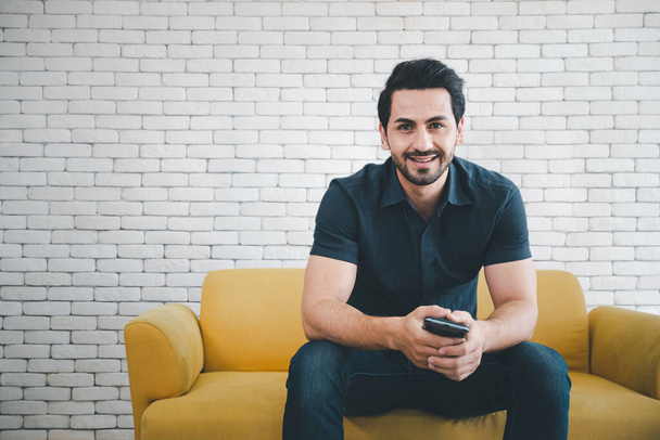 happy man with smiling face sitting on yellow sofa with smartphone looking at camera, lifestyle concept - Foto, imagen