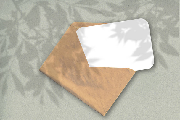 Un sobre con una hoja de papel blanco texturizado sobre el fondo gris de la pared. Mockup superpuesto con las sombras de la planta. La luz natural proyecta sombras desde el follaje de los árboles - Foto, imagen