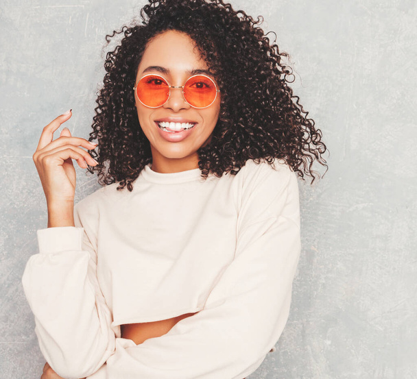 Beautiful black woman with afro curls hairstyle.Smiling model in white trendy jeans clothes and sunglasses. Sexy carefree female posing near gray wall in studio. Tanned and cheerful - Photo, Image