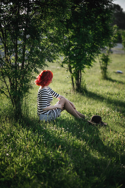 Beautiful landscape with a young girl sitting under a tree. A hipster girl with red hair sits on the grass in thought. Relaxation, enjoying nature, traveling. - Fotografie, Obrázek