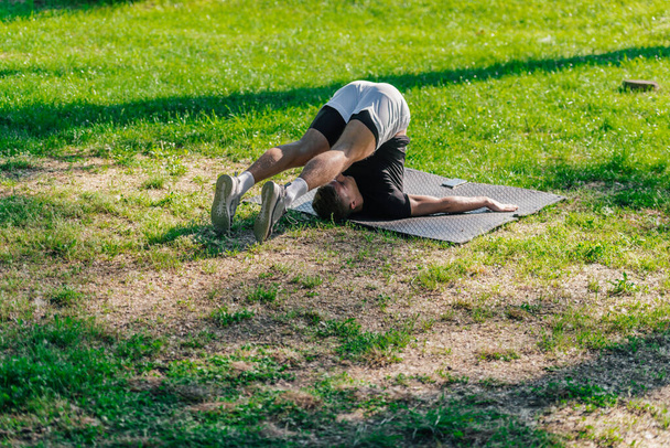 Mladý sportovní pohledný muž cvičit jógu v parku a dělat pluhu pózu. - Fotografie, Obrázek