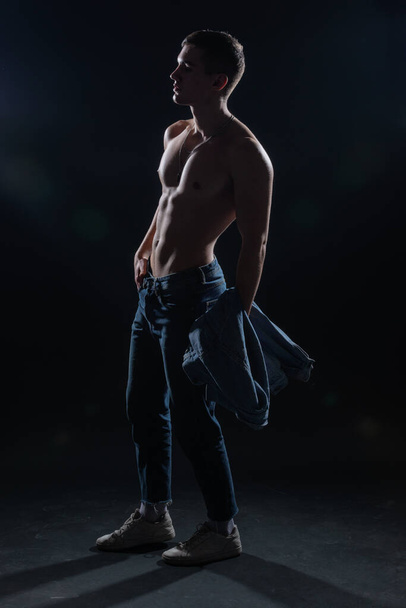 Sylwetka gorącego muskularnego mężczyzny pozującego w dżinsach w ciemnym studiu. - Zdjęcie, obraz