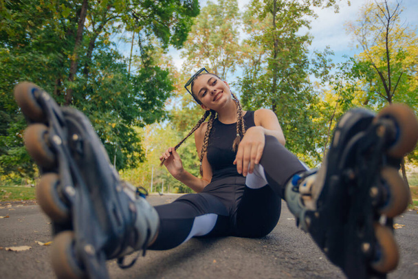 Verbazingwekkende jonge vrouw met haar vlechten poseren in rolschaatsen terwijl zitten op een weg in het park - Foto, afbeelding