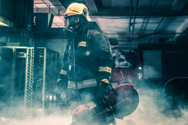 Портрет пожежника, який носить повне захисне обладнання, позує з бензопилою на плечі. Темний фон з димом і синім світлом
. - Фото, зображення