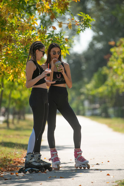 Deux belles meilleures amies parlent et boivent des smoothies dans le parc tout en se tenant debout sur leurs patins à roulettes - Photo, image