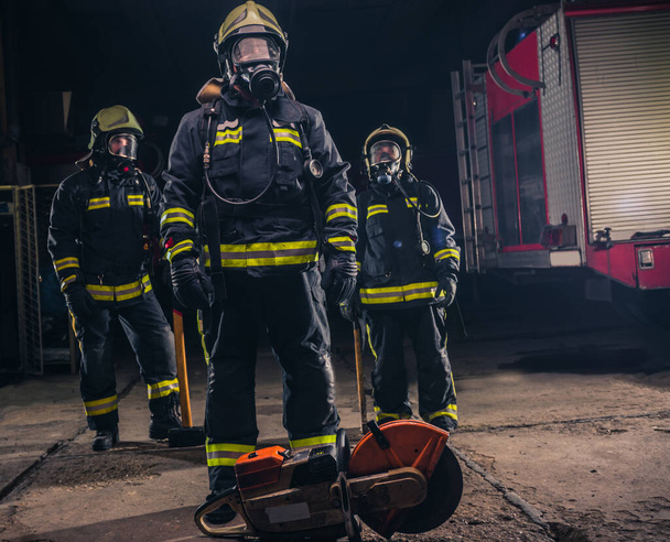 Grupo de tres jóvenes bomberos posando dentro del departamento de bomberos con uniforme y herramientas - Foto, imagen