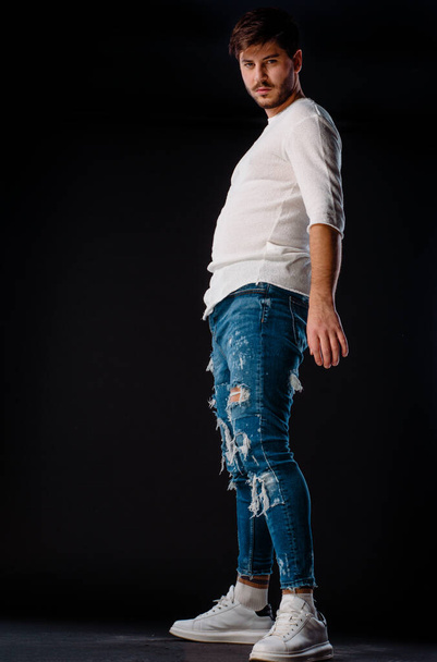 Schöner kleiner Junge trägt Hipster-Jeans, während er vor dunklem Hintergrund posiert - Foto, Bild