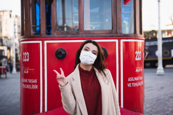 Hermosa chica con máscara médica protectora y ropa de moda posa con tranvía rojo en la calle istiklal en Estambul, Turquía. Nuevo concepto de estilo de vida normal. - Foto, imagen