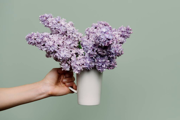 Lilac flowers in white cup in the hands of woman on green background - Foto, Imagen