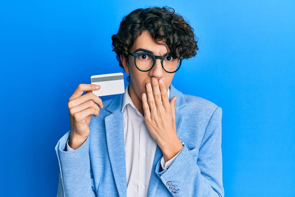 Latino nuori mies, jolla luottokortti peittää suun käsin, järkyttynyt ja pelkää erehdystä. yllättynyt ilme  - Valokuva, kuva
