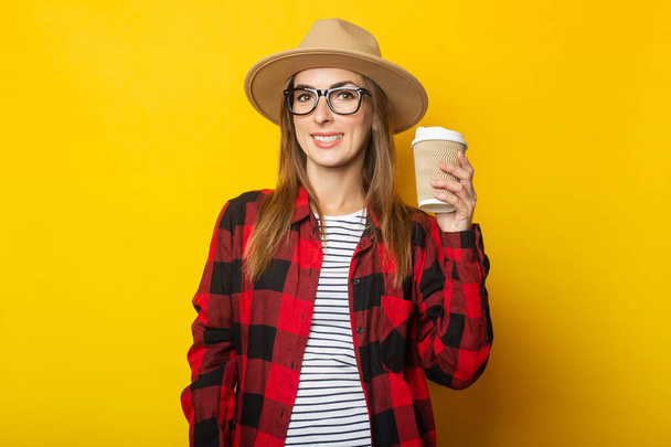 Şapkalı ve ekose gömlekli genç bir kadın elinde kahve ve sarı arka planda bir kağıt bardak tutuyor.. - Fotoğraf, Görsel