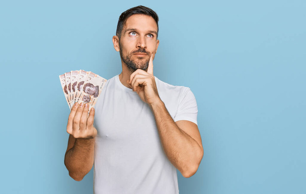 Schöner Mann mit Bart, der 50 mexikanische Pesos-Banknoten hält, ernstes Gesicht, das über Fragen nachdenkt, mit der Hand am Kinn, nachdenklich über verwirrende Idee  - Foto, Bild