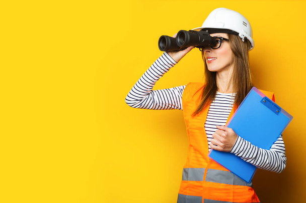 Jovem mulher em colete de sinal e capacete de construção olhando através de binóculos no fundo amarelo. - Foto, Imagem