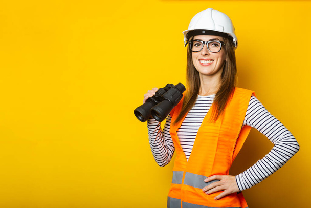 Young woman with a smile in a vest and a helmet holds binoculars on a yellow background. Construction concept, new building. Banner. - Photo, Image