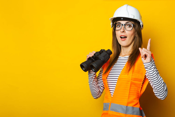 Young woman with a surprised face in a vest and hard hat holds binoculars on a yellow background. Construction concept, new building. Banner. - Photo, image