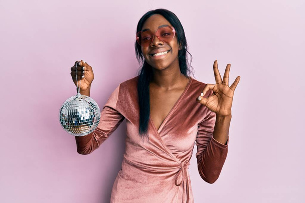 Seksi parti elbisesi giymiş, elinde disko topu tutan genç Afrikalı Amerikalı kadın parmaklarıyla imza atıp gülümseyerek mükemmel bir sembol oluşturuyor.  - Fotoğraf, Görsel