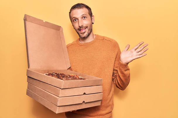 Young handsome man holding delivery pizza boxes celebrating achievement with happy smile and winner expression with raised hand  - Photo, Image