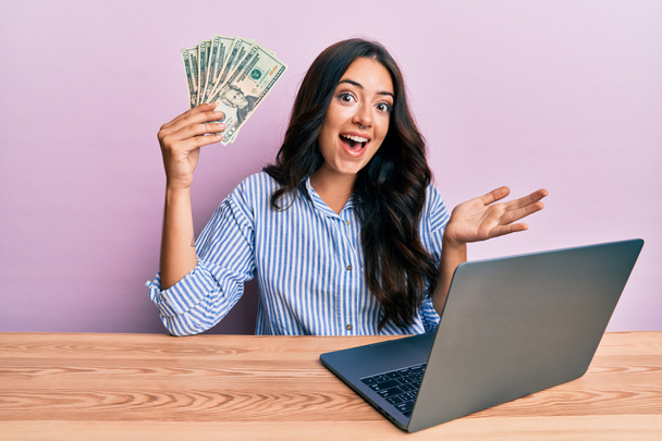 Красивая брюнетка молодая женщина, работающая в офисе, держа в руках доллары, празднуя достижение с счастливой улыбкой и выражением победителя с поднятой рукой  - Фото, изображение