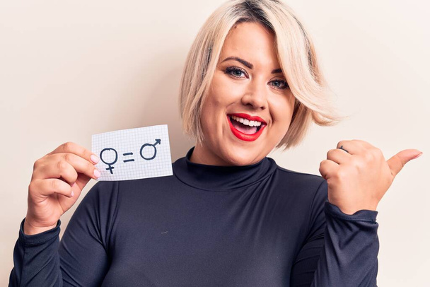Blonde plus size woman asking for sex equality holding paper with gender equal message pointing thumb up to the side smiling happy with open mouth - Photo, Image
