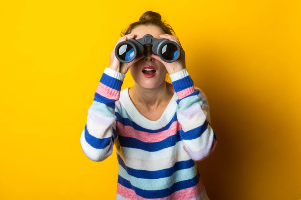 Mujer joven en suéter mirando a través de binoculares sobre fondo amarillo. - Foto, imagen