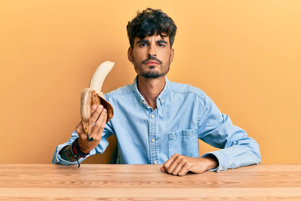 Young hispanic man sitting on the table holding banana thinking attitude and sober expression looking self confident  - Photo, Image
