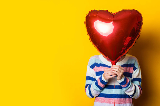 Молодая женщина в свитере прячется за сердцем воздушного шара на жёлтом фоне. Сочинение ко Дню Святого Валентина. - Фото, изображение
