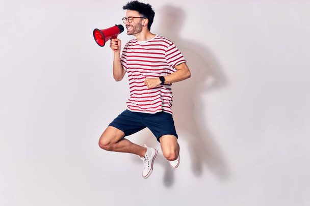 Young handsome hispanic man wearing glasses screaming using megaphone. Jumping over isolated white background - Foto, Bild