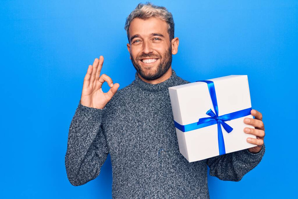 Junger schöner blonder Mann mit Bart hält Geburtstagsgeschenk über isoliertem blauem Hintergrund und tut ok Zeichen mit den Fingern, lächelt freundlich gestikulierend hervorragendes Symbol - Foto, Bild