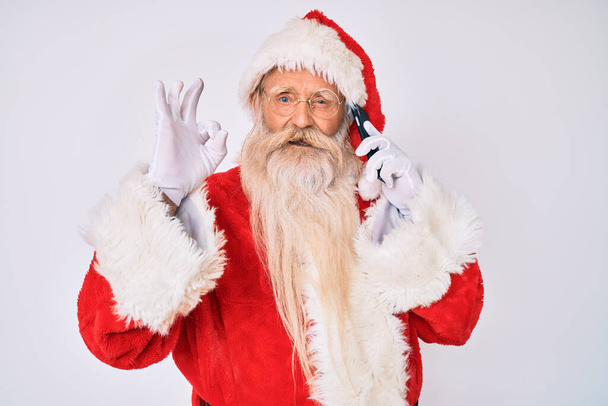 Old senior man wearing santa claus costume speaking on the phone doing ok sign with fingers, smiling friendly gesturing excellent symbol  - Fotoğraf, Görsel