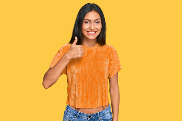 Jeune femme arabe brune portant des vêtements décontractés faisant des pouces heureux geste avec la main. approuvant l'expression regardant la caméra montrant le succès.  - Photo, image