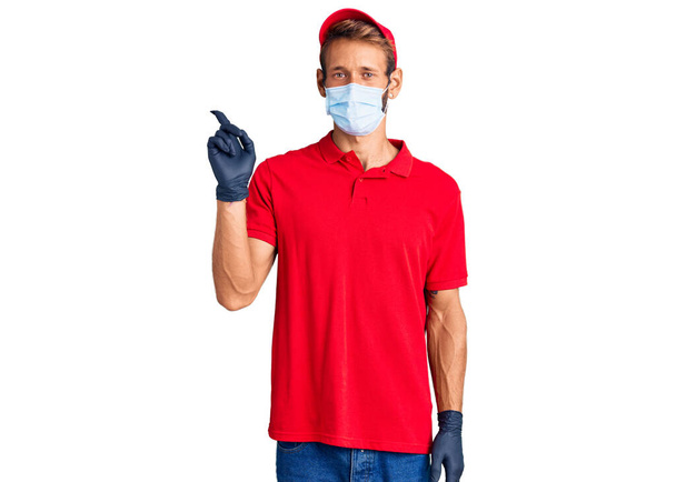 Handsome blond man with beard wearing delivery uniform and medical mask smiling happy pointing with hand and finger to the side  - Photo, Image