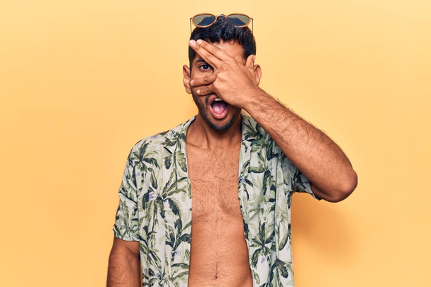 Young latin man wearing summer shirt and sunglasses peeking in shock covering face and eyes with hand, looking through fingers afraid  - Fotografie, Obrázek