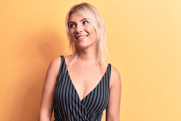 Young beautiful blonde woman wearing striped t-shirt standing over isolated yellow background looking to side, relax profile pose with natural face and confident smile. - Foto, immagini
