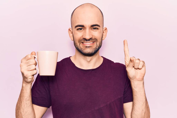 Junger gutaussehender, glatzköpfiger Mann, der eine Tasse Kaffee trinkt, mit einer Idee oder Frage lächelnd mit glücklichem Gesicht zeigt, Nummer eins  - Foto, Bild
