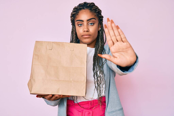 Νεαρή αφροαμερικανίδα επιχειρηματίας με πλεξούδες κρατώντας πάρει μακριά χαρτοσακούλα με ανοιχτό χέρι κάνει στοπ σημάδι με σοβαρή και σίγουρη έκφραση, χειρονομία άμυνας  - Φωτογραφία, εικόνα