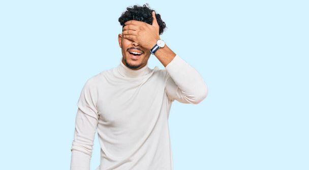 Jonge Arabier man draagt casual winter trui glimlachend en lachend met de hand op het gezicht bedekt ogen voor verrassing. blind concept.  - Foto, afbeelding