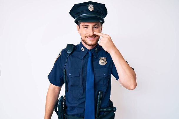 Junger kaukasischer Mann in Polizeiuniform, der mit der Hand auf Gesicht und Nase zeigt und fröhlich lächelt. Schönheitskonzept  - Foto, Bild