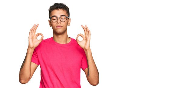 カジュアルな服を着た若いハンサムなアフリカ系アメリカ人男性は、指で瞑想ジェスチャーを行う閉じて目でリラックスして笑顔。ヨガのコンセプト.  - 写真・画像