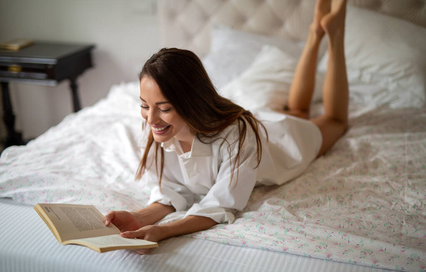 Rest, comfort, leisure and people concept. Sexy happy woman reading book in bed at home - Zdjęcie, obraz