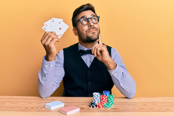 Красивый латиноамериканец-крупье, сидящий на столе с фишками для покера и серьезными картами, думающий о вопросе с подбородком, думающий о запутанной идее  - Фото, изображение