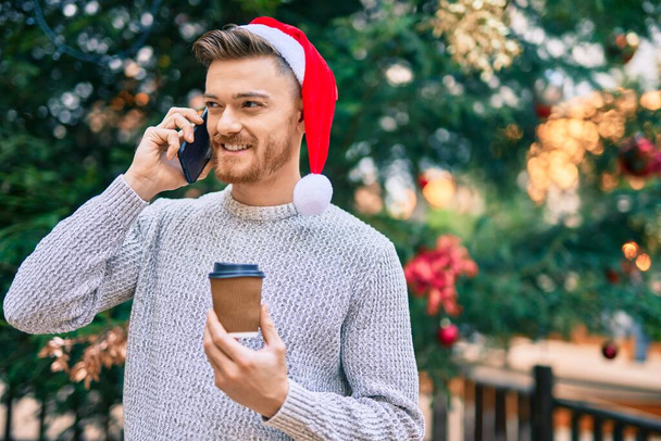 Junger Kaukasier mit Weihnachtsmütze plaudert auf dem Smartphone und trinkt Kaffee im Park - Foto, Bild