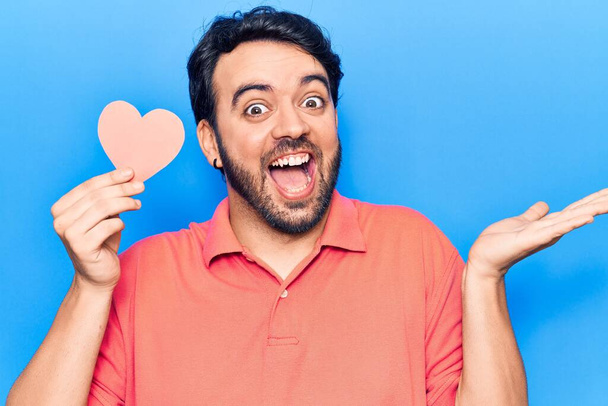 Young hispanic man holding heart shape paper celebrating achievement with happy smile and winner expression with raised hand  - Foto, imagen