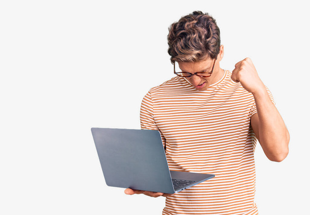 Young handsome man wearing glasses holding laptop annoyed and frustrated shouting with anger, yelling crazy with anger and hand raised  - Fotoğraf, Görsel